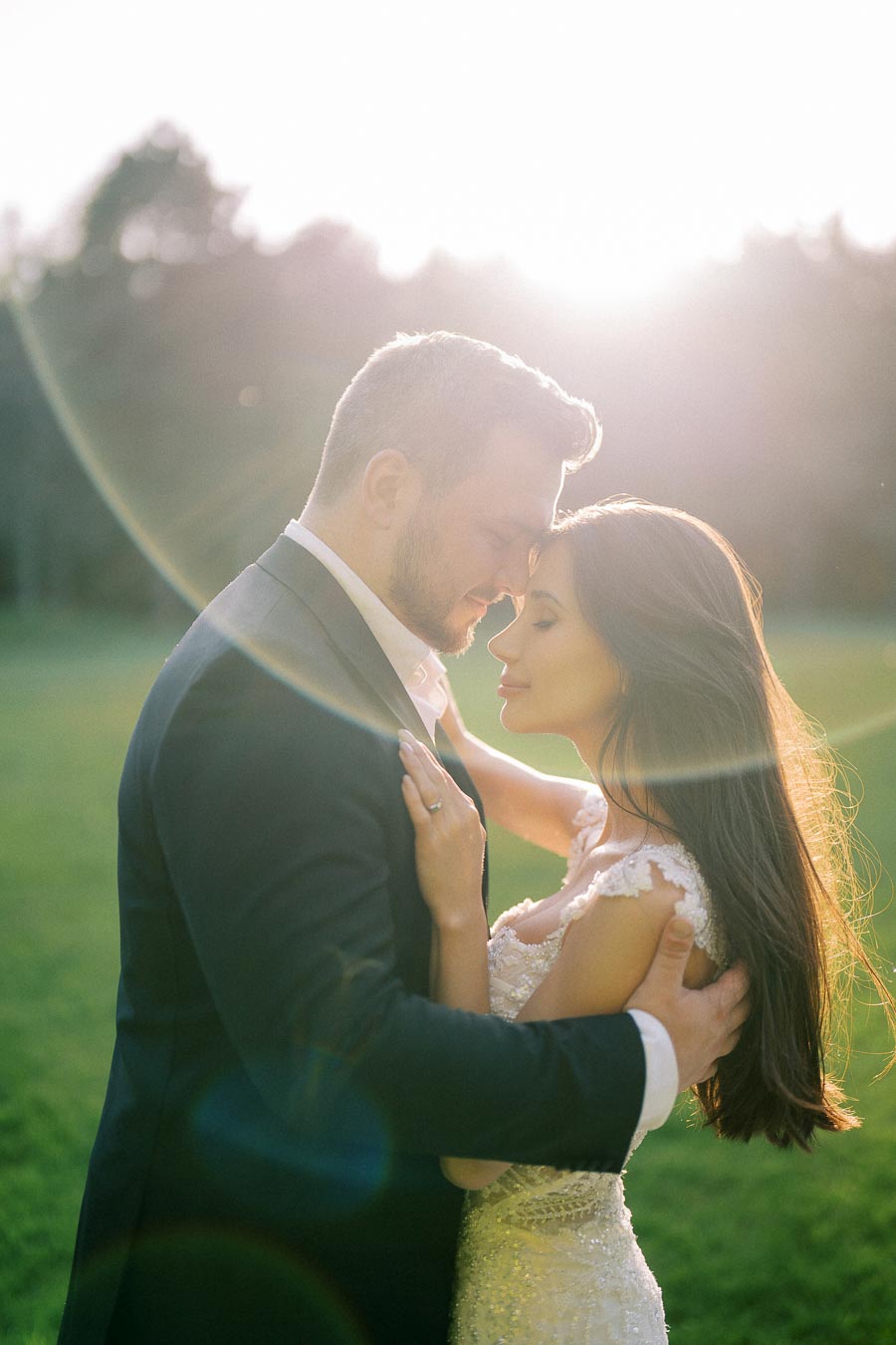 A romantic couple embraces outdoors at sunset, bathed in warm sunlight. The groom is in a dark suit, and the bride is wearing a lacy white dress, standing in a lush green field, with soft lens flare creating a dreamy effect.