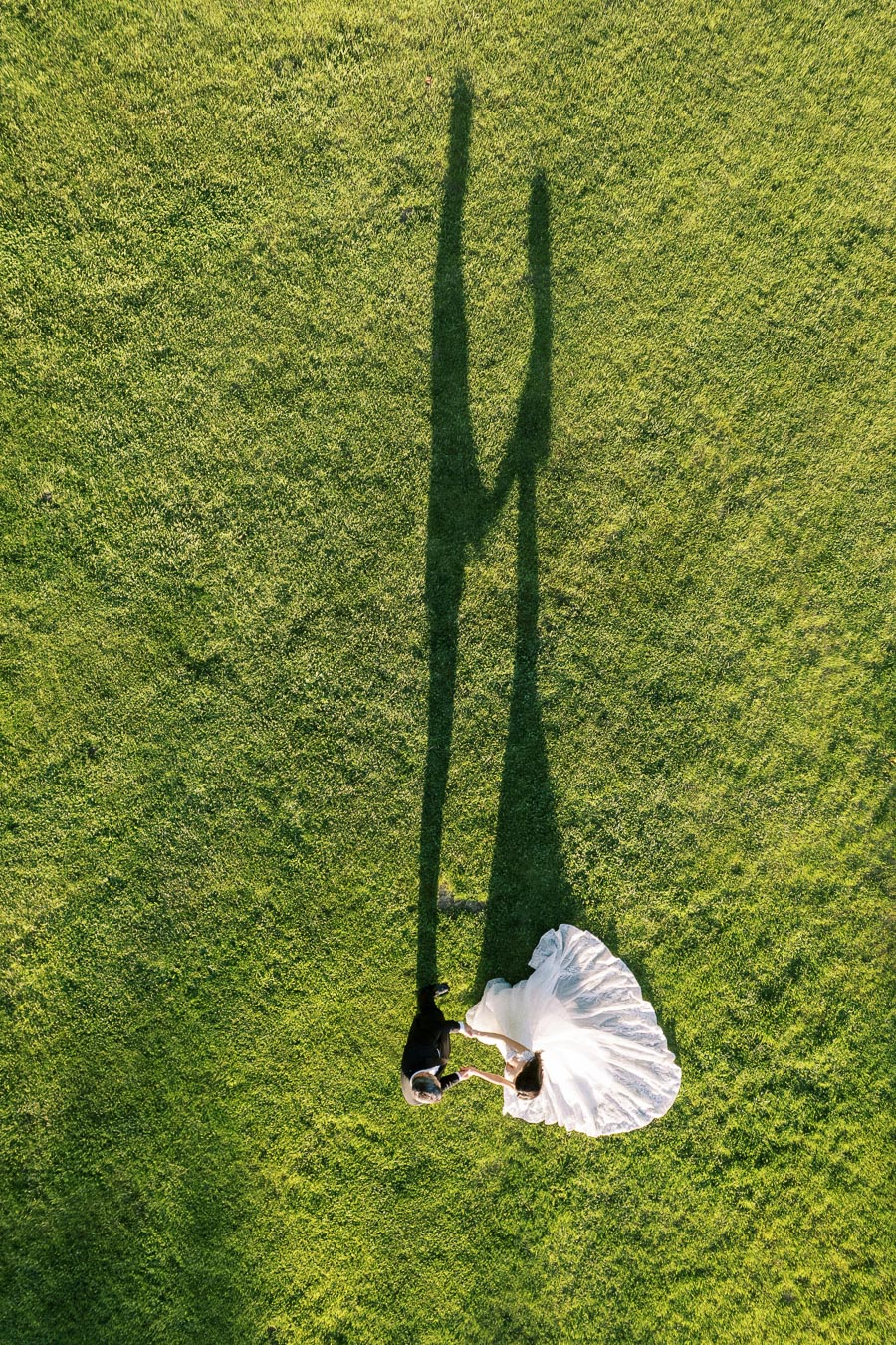 Aerial view of a couple dancing on a grassy field, casting long shadows in the sunlight, with the bride in a flowing white dress and the groom in a black suit.