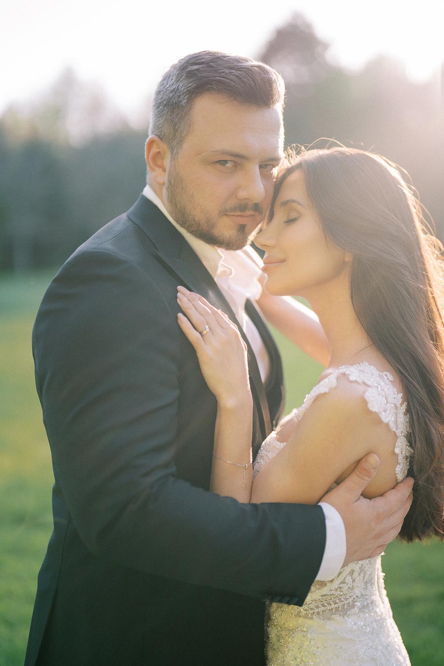 Romantic couple embracing in a sunlit outdoor setting, man wearing a dark suit and woman in an elegant lace wedding dress, with serene expressions.
