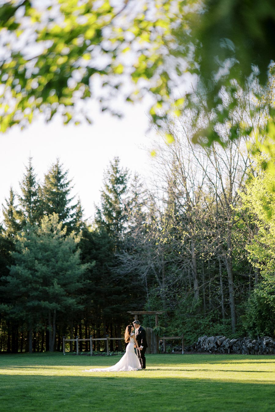 Romantic outdoor wedding scene with bride and groom standing on lush green grass, surrounded by scenic forest backdrop under a canopy of leaves.