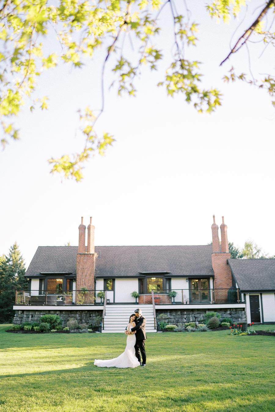A bride and groom embrace on a lush green lawn in front of a charming house with brick chimneys, surrounded by trees under a clear sky.
