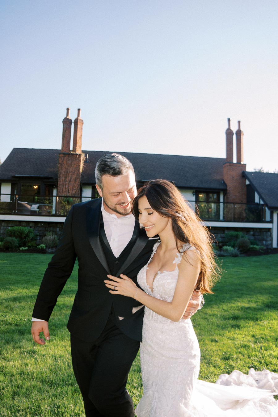 A joyful bride and groom, in a white lace wedding dress and black tuxedo, walking hand in hand on a sunny day in front of a charming house with a green lawn.