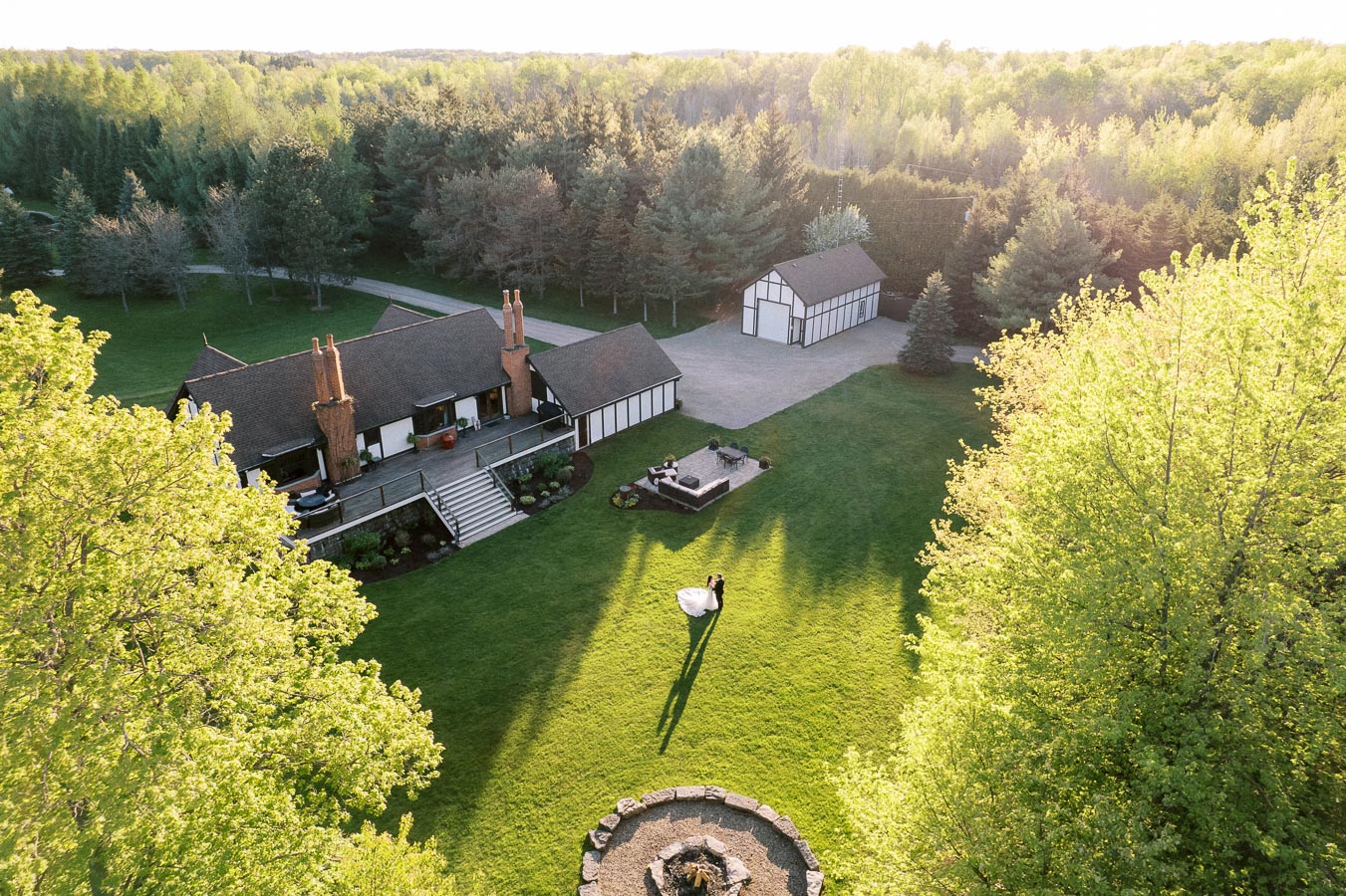 Aerial view of a countryside estate featuring a large house and outbuilding surrounded by lush greenery and trees, with a couple dancing on the expansive lawn under the golden hour sunlight.