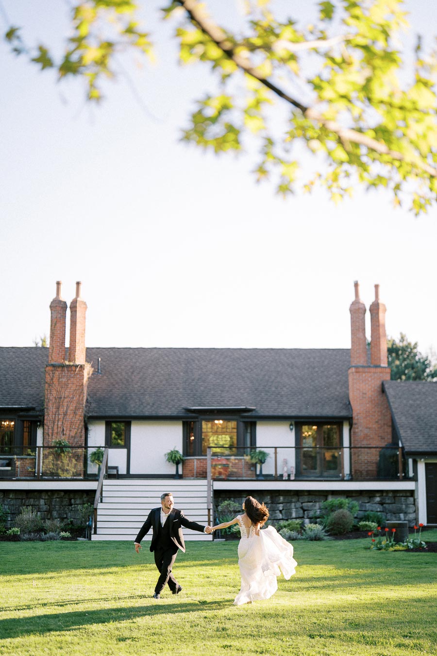 A bride and groom joyfully running on a lush green lawn in front of a charming house with a brick chimney, surrounded by vibrant landscaping and under a clear blue sky with sunlit leaves in the foreground.