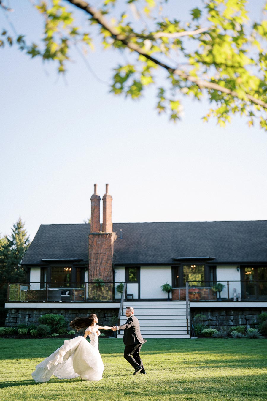 A bride and groom joyfully dance on a lush green lawn in front of a charming house, with sunlight filtering through the leafy branches above.