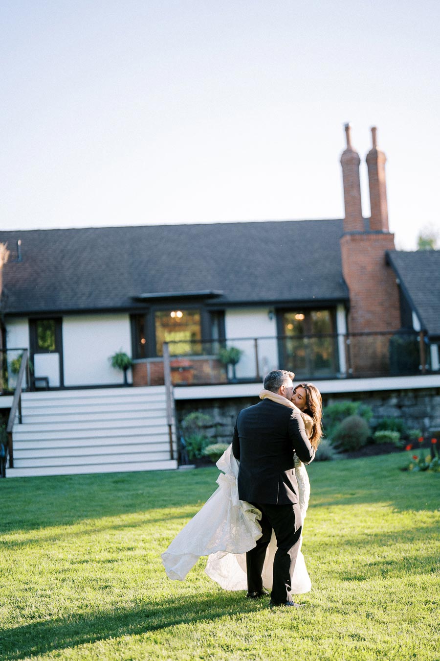 A couple in formal attire embraces in front of a charming house with large brick chimneys, standing on a lush green lawn under a clear blue sky.
