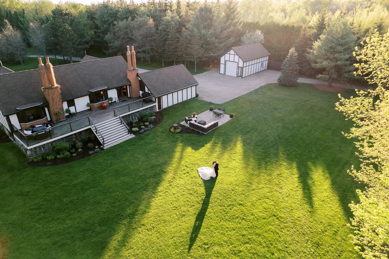 Aerial view of a scenic rustic house surrounded by lush green trees and a well-maintained lawn, with a couple sharing a romantic moment in the garden, casting long shadows in the evening light.