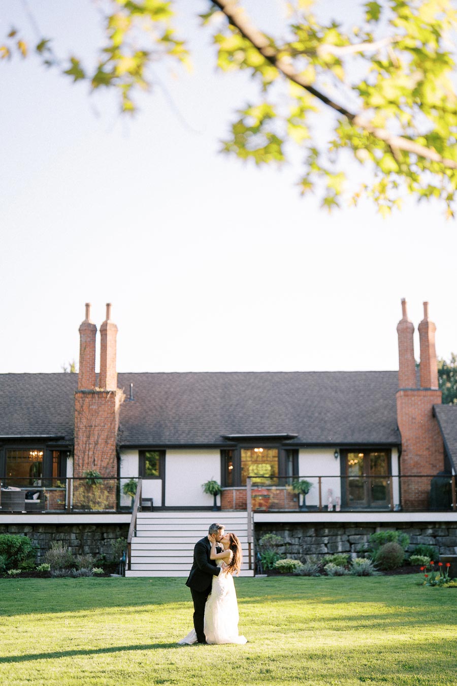 A bride and groom share a romantic kiss on a lush green lawn in front of a charming cottage-style house with brick chimneys, under a clear blue sky and beside vibrant foliage.