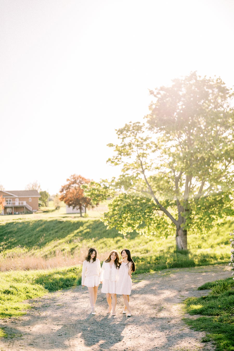 Three women in white dresses walking together on a sunny path through a lush green landscape, highlighting friendship and nature's beauty.