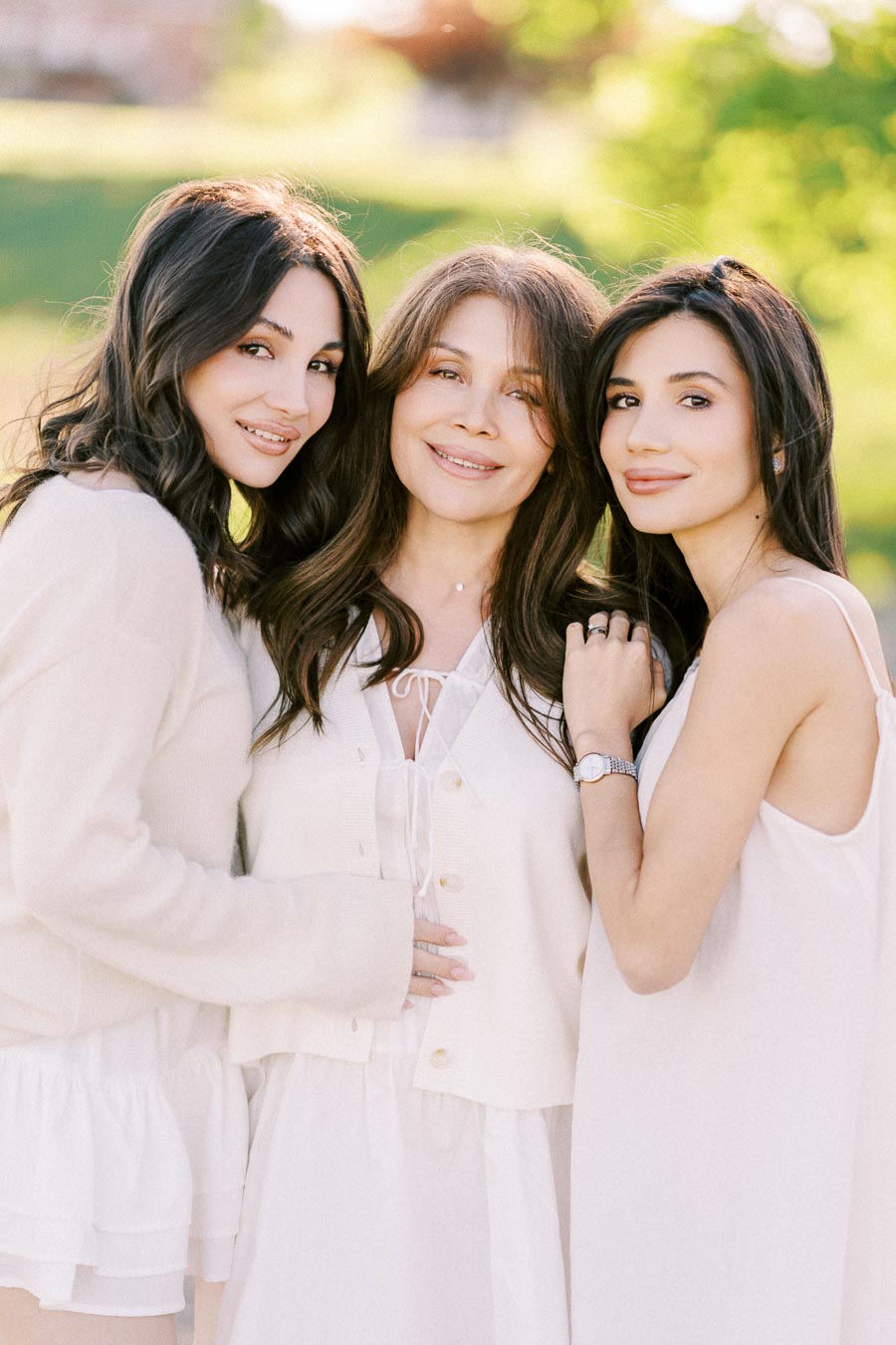 A group of three women in white dresses posing together outdoors with a blurred green background, emphasizing a sense of family and happiness.
