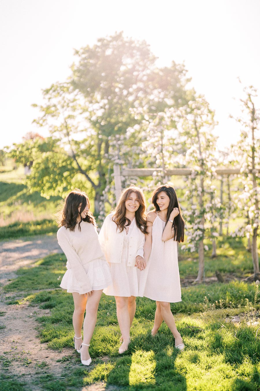 Three women in white dresses enjoying a sunny day in a lush green orchard