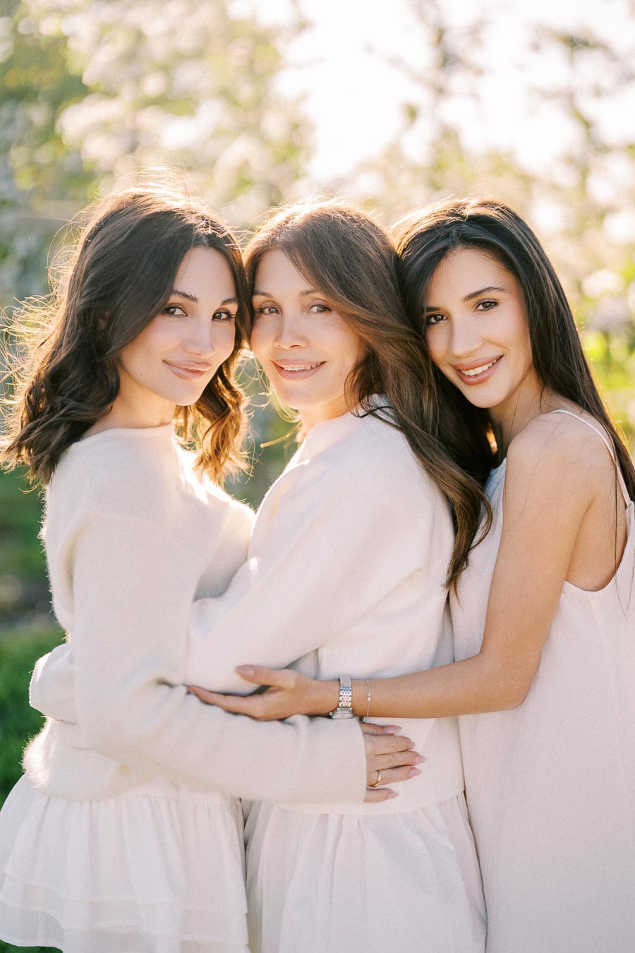 Three smiling women in light-colored dresses embracing each other outdoors with a sunlit backdrop.