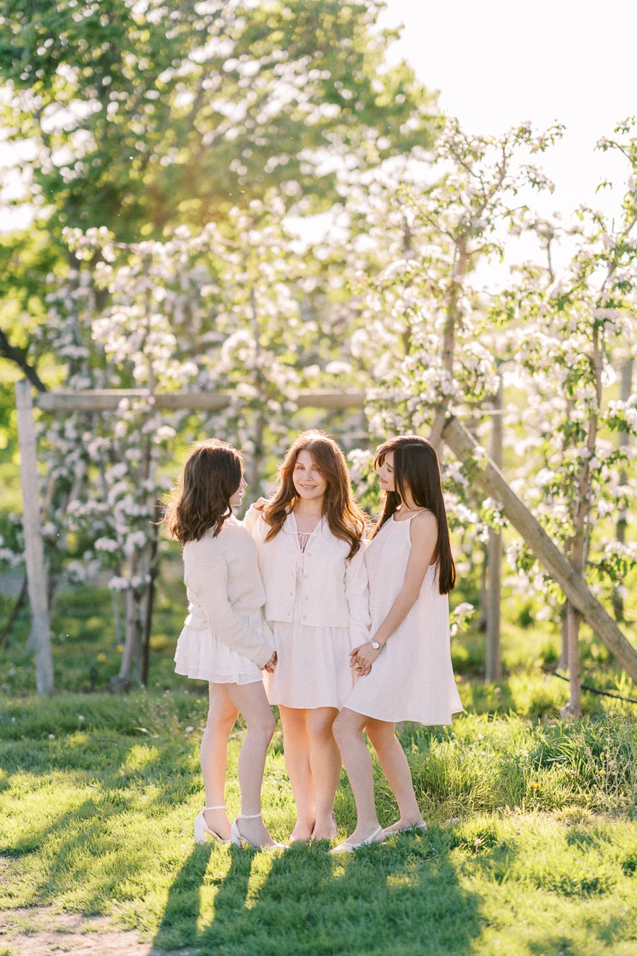 Three women in light dresses standing together in a sunlit orchard with blossoming trees.