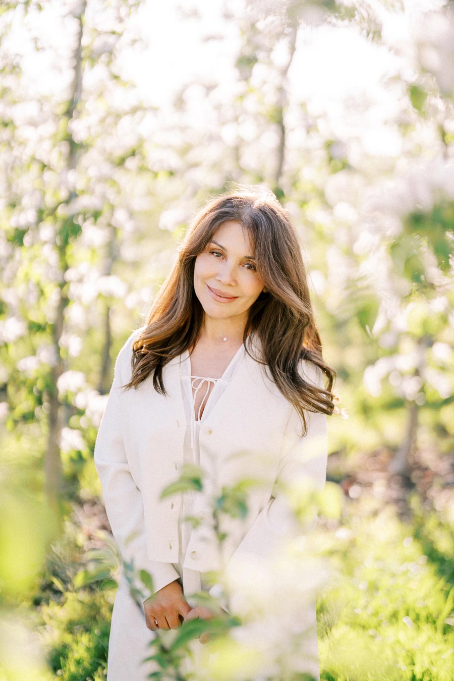 A woman in a white jacket, standing confidently in a sunlit garden with blooming trees, smiling at the camera.