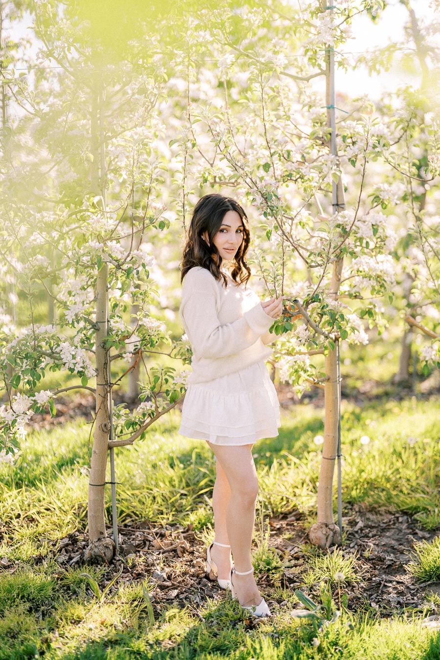 Young woman in a white dress standing in a sunlit orchard, surrounded by blooming trees and fresh green grass.