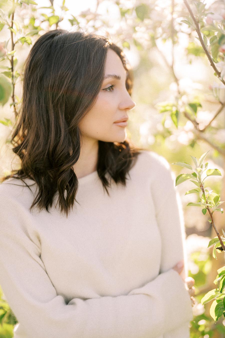 A woman in a cream sweater stands amidst blooming trees, with sunlight filtering through the branches, creating a serene and natural atmosphere.