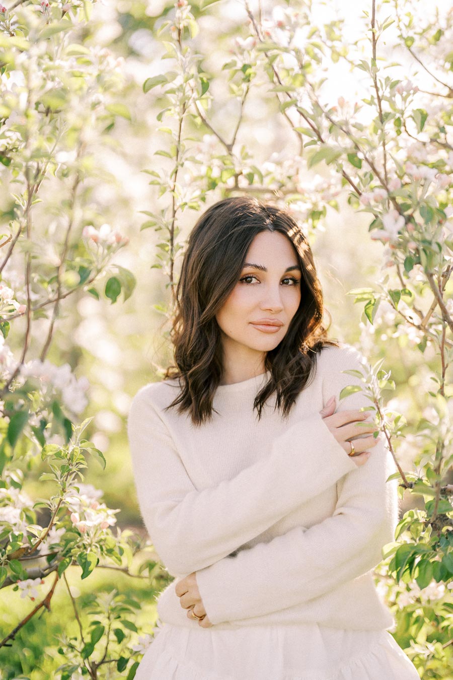 Woman in a white sweater standing amidst blooming trees in a garden, with sunlight filtering through the leaves, creating a serene and natural atmosphere.
