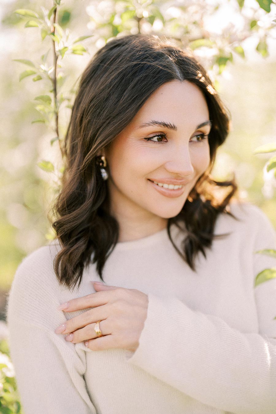 A smiling woman with dark hair wearing a white sweater stands in a sunlit garden, surrounded by green leaves, showcasing natural beauty and outdoor serenity.