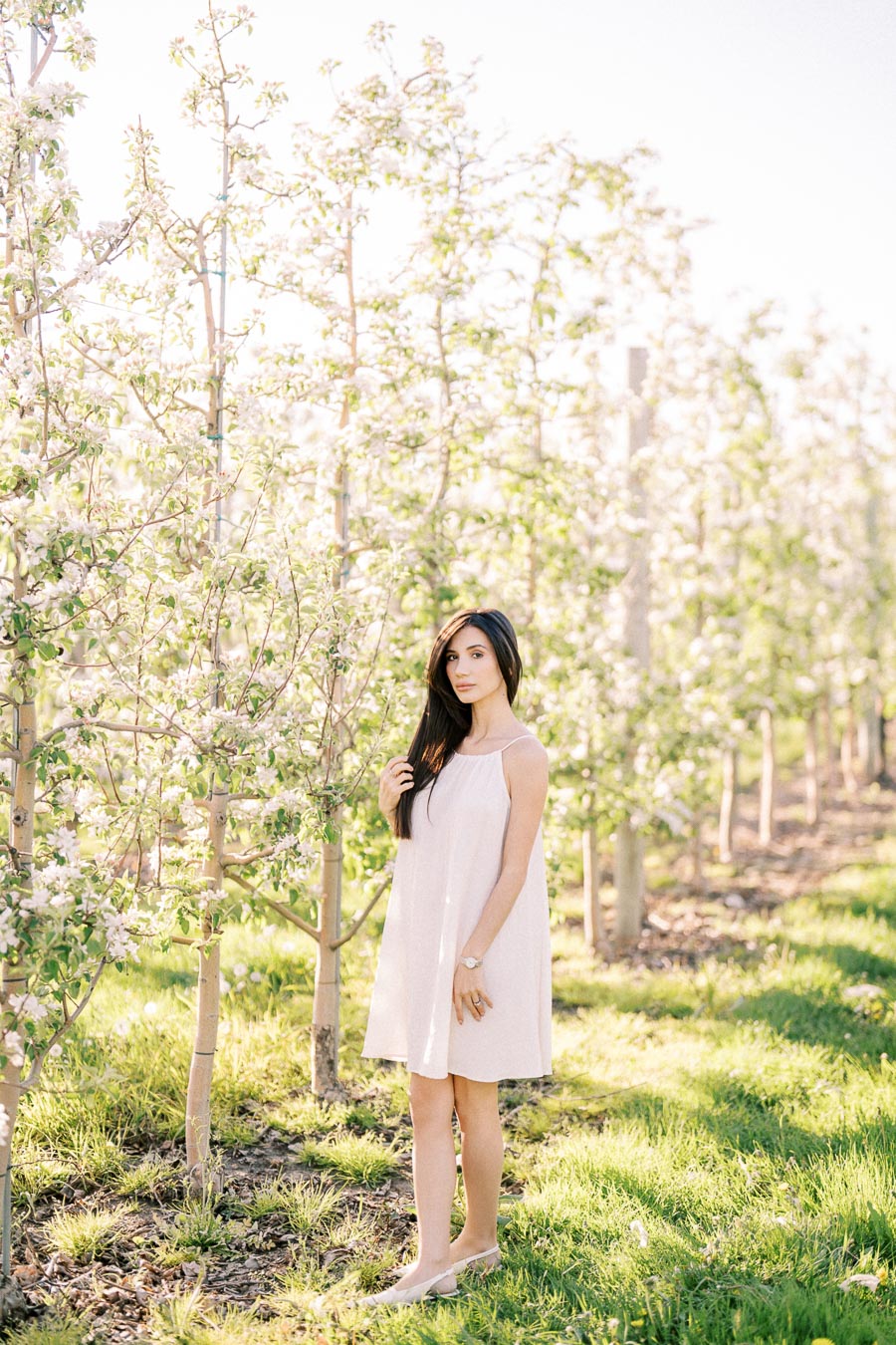 A person in a white dress standing in a lush orchard with blooming trees under a clear sky.