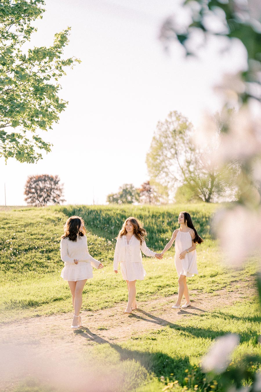 Three young women walking hand in hand through a sunny, grassy field, surrounded by lush greenery and blossoms, wearing white dresses and enjoying a bright spring day.