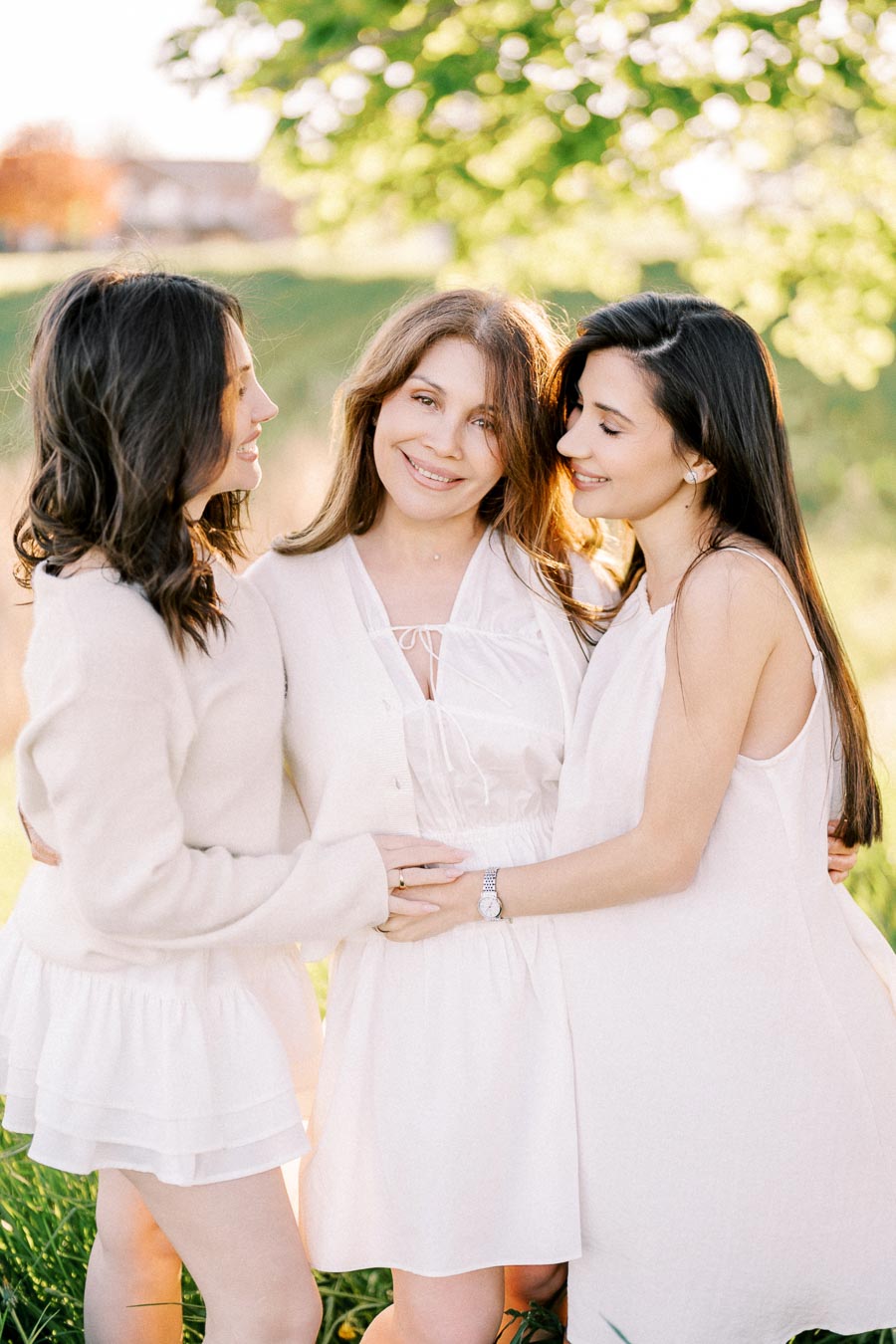 Three women in white dresses smiling and embracing outdoors on a sunny day, with a blurred green leafy background, showcasing friendship and joy.