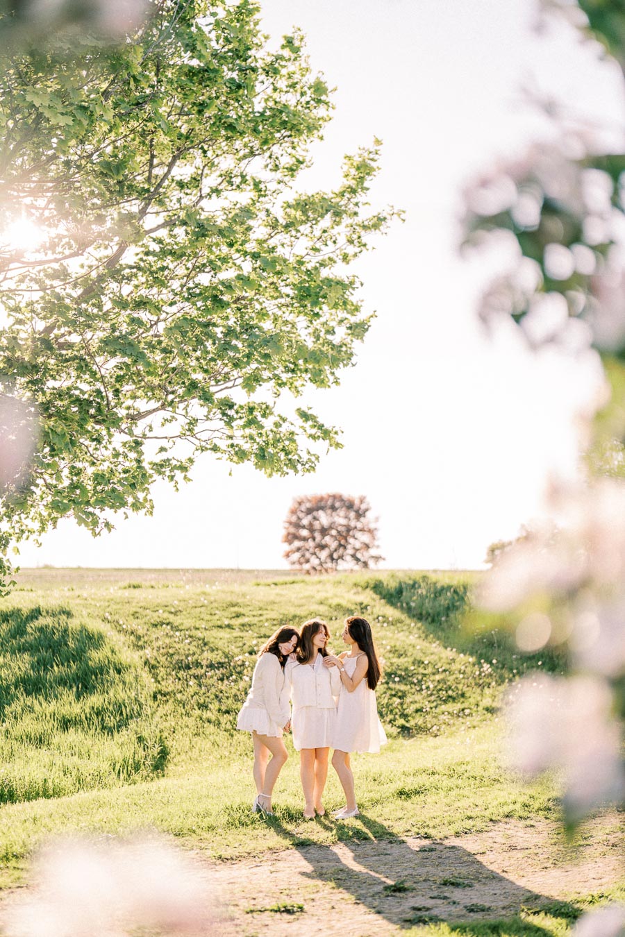 Three women in white dresses standing in a sunlit, grassy field, surrounded by trees and flowers, enjoying a spring day together.