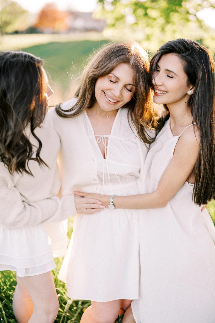 Three women in white dresses enjoying a sunny day outdoors while embracing each other and smiling, surrounded by lush greenery.
