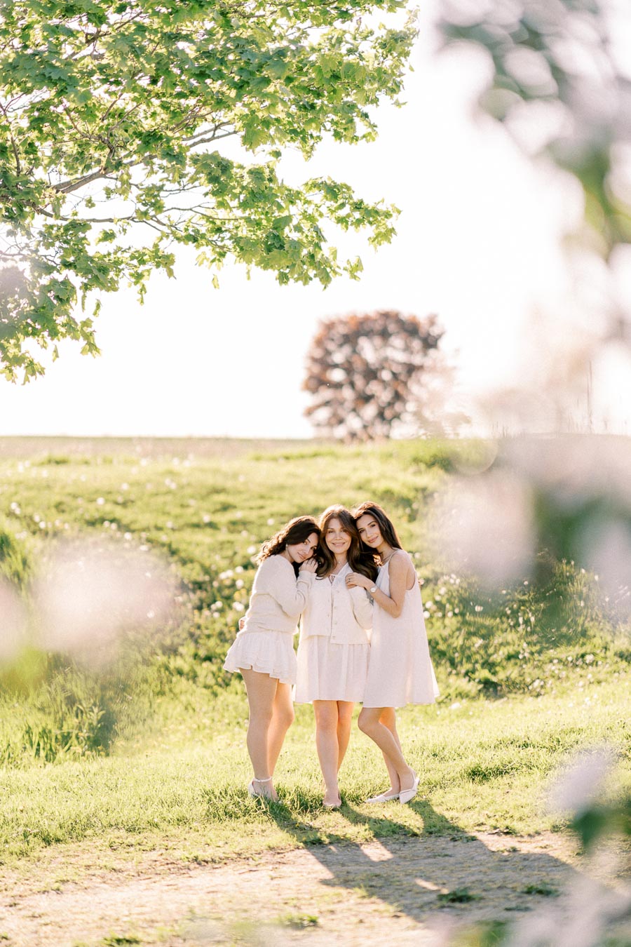 Three women in white dresses standing together in a sunny, green field with trees in the background, capturing a peaceful outdoor moment.