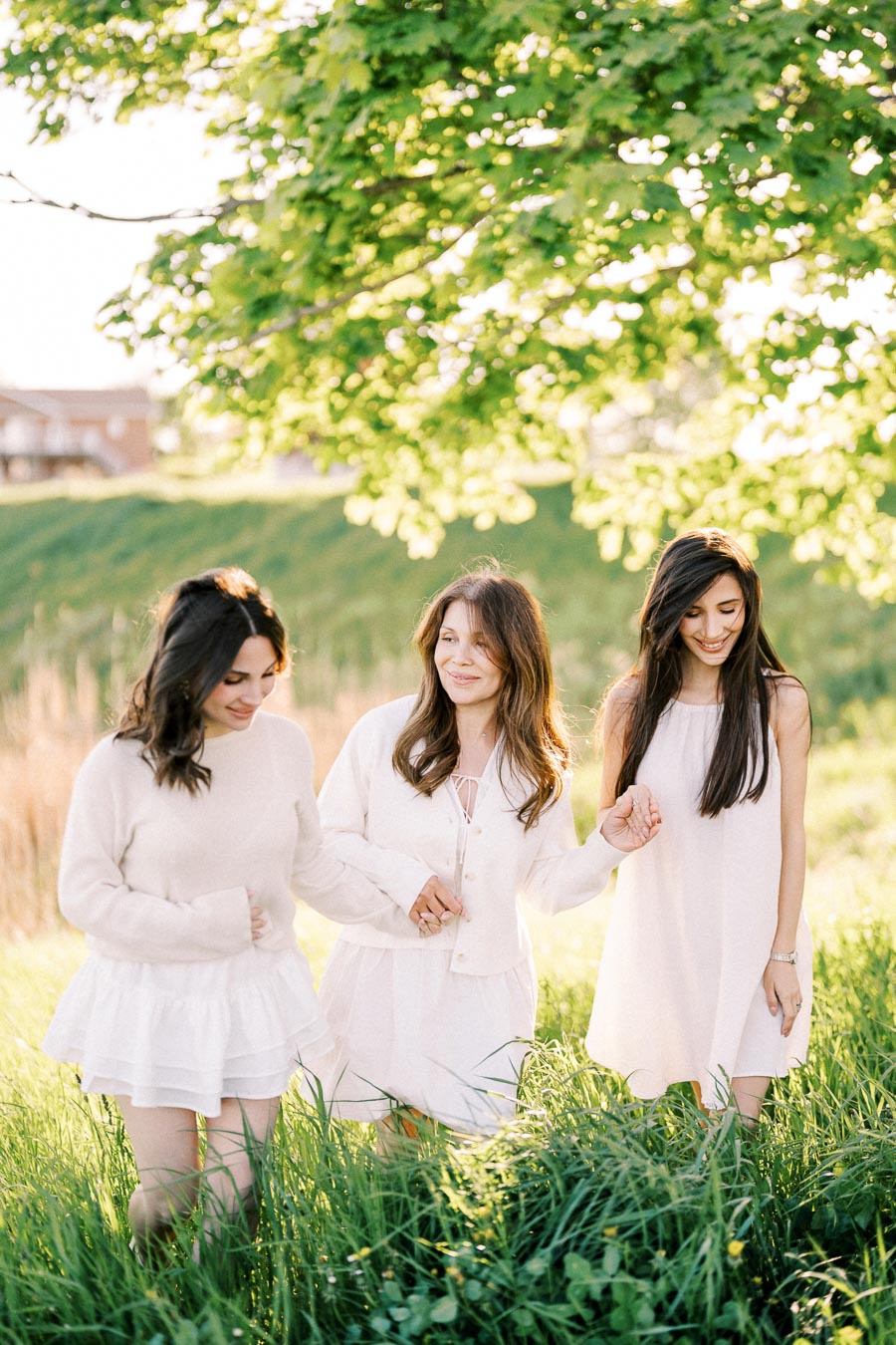 Three women in white dresses walking hand in hand through a sunny, grassy field with lush green trees in the background.