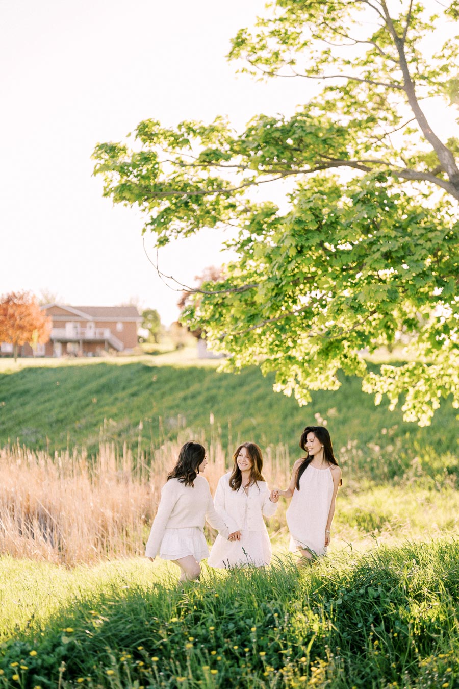 Three women enjoying a sunny day in a grassy field under a tree, dressed in light spring clothing.