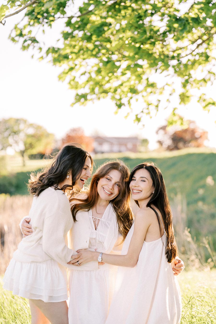 Three women in white dresses, smiling and embracing under a tree on a sunny day. They are standing in a grassy field, with a house visible in the background.