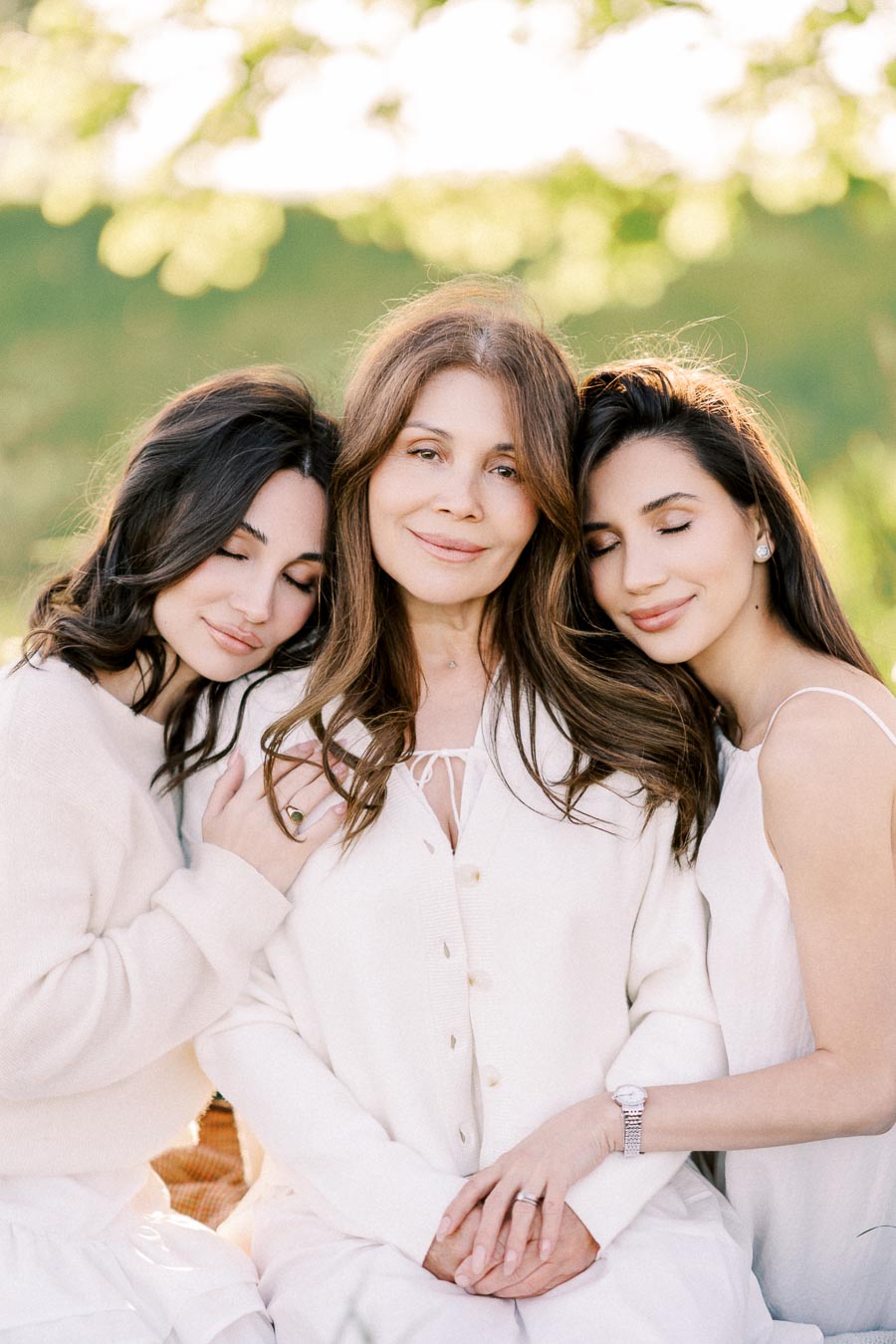 A mother and two daughters embracing each other in a serene outdoor setting, wearing white outfits, expressing warmth and family bonding.