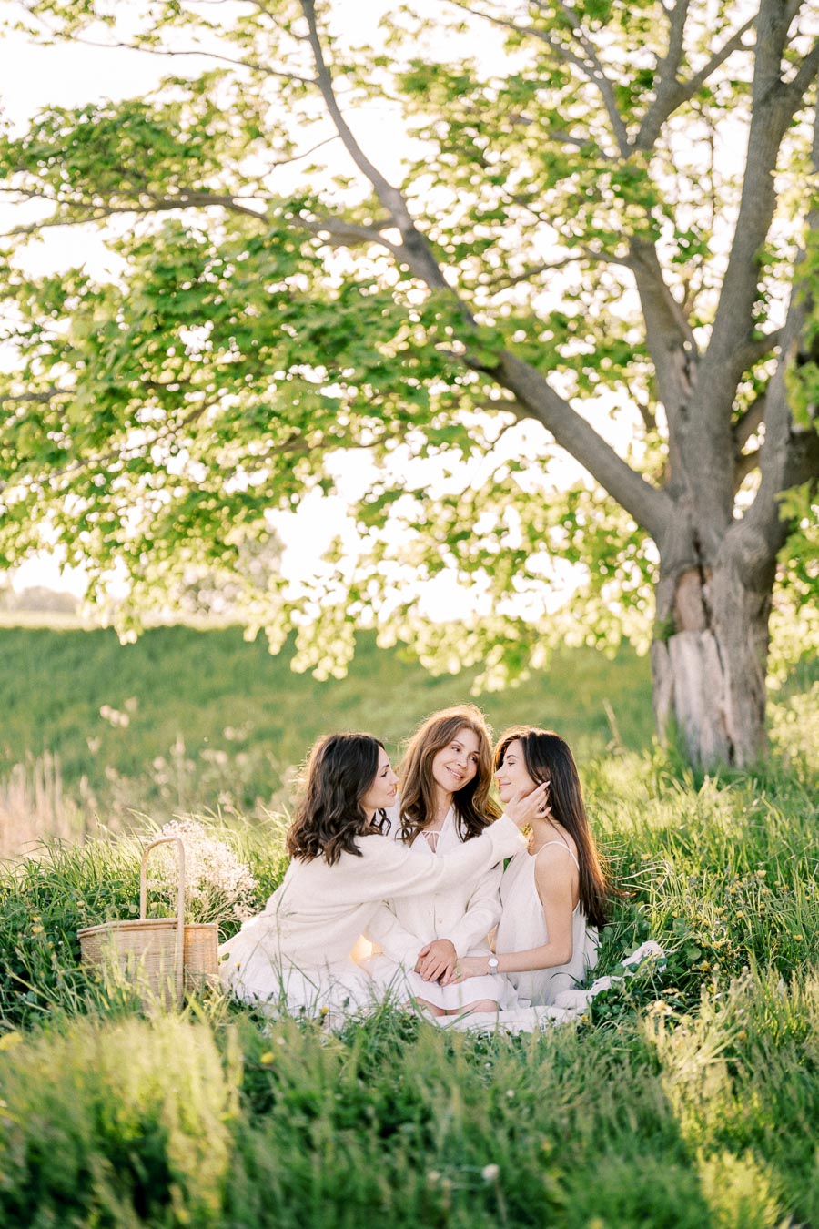 Three women enjoying a picnic in a lush green meadow under a large tree, surrounded by sunlight and nature.