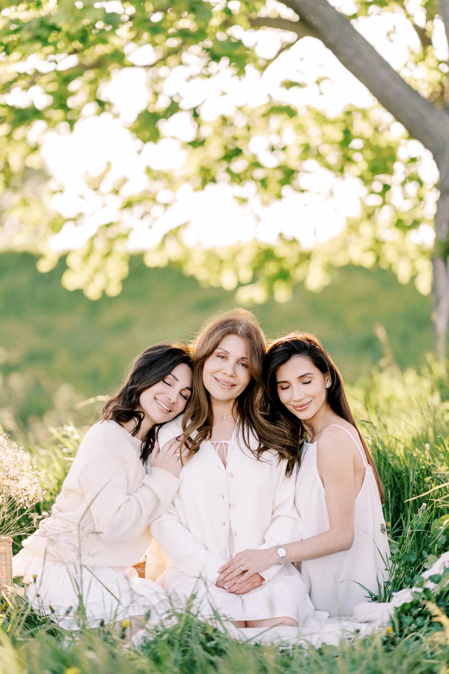 Three women sitting together in a lush green field, wearing coordinated light-colored outfits, smiling and enjoying a sunny day under a tree. The background features vibrant foliage, creating a serene and joyful atmosphere.