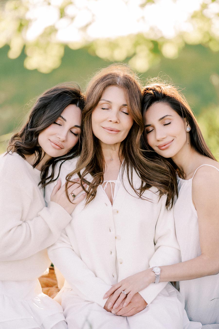 Three women in white clothing sitting closely together outdoors, with eyes closed, embracing and enjoying a serene moment in a sunlit, natural setting.
