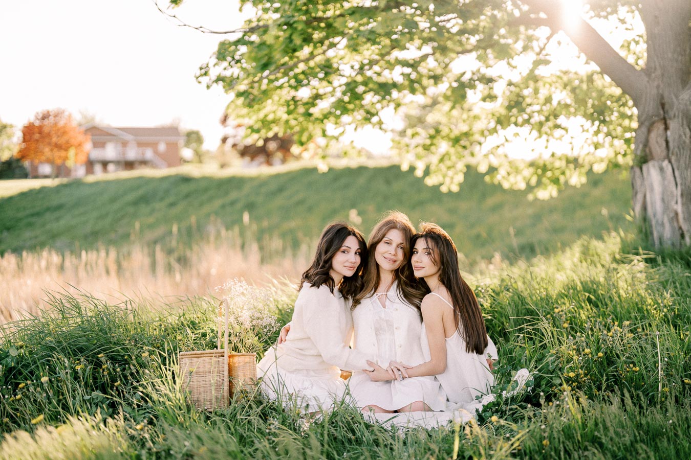 A serene outdoor scene featuring three women sitting on lush green grass under a large tree, enjoying a sunny afternoon. They are dressed in light-colored clothing, with a wicker basket nearby, creating a peaceful and harmonious atmosphere suitable for a spring or summer picnic.