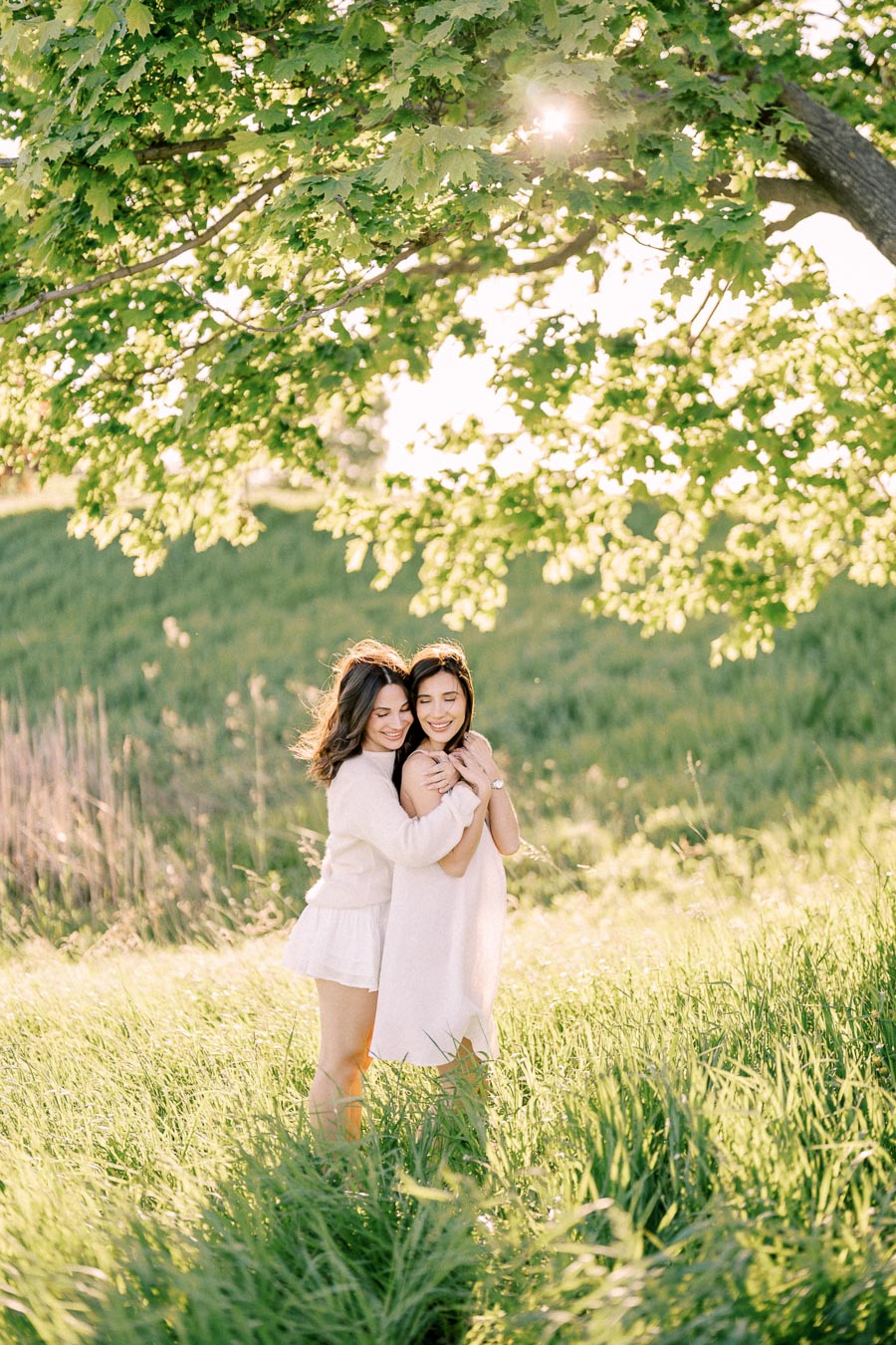 Two women embracing under a sunlit tree in a lush green field, with sunlight filtering through the leaves.