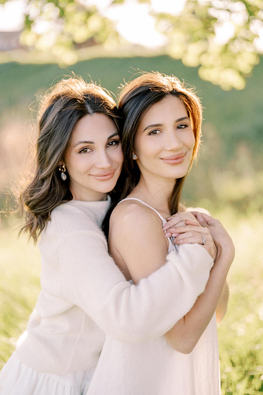 Two women embracing in a sunlit outdoor setting, smiling warmly at the camera. They are standing in a grassy field with blurred greenery in the background, conveying a sense of warmth and closeness.