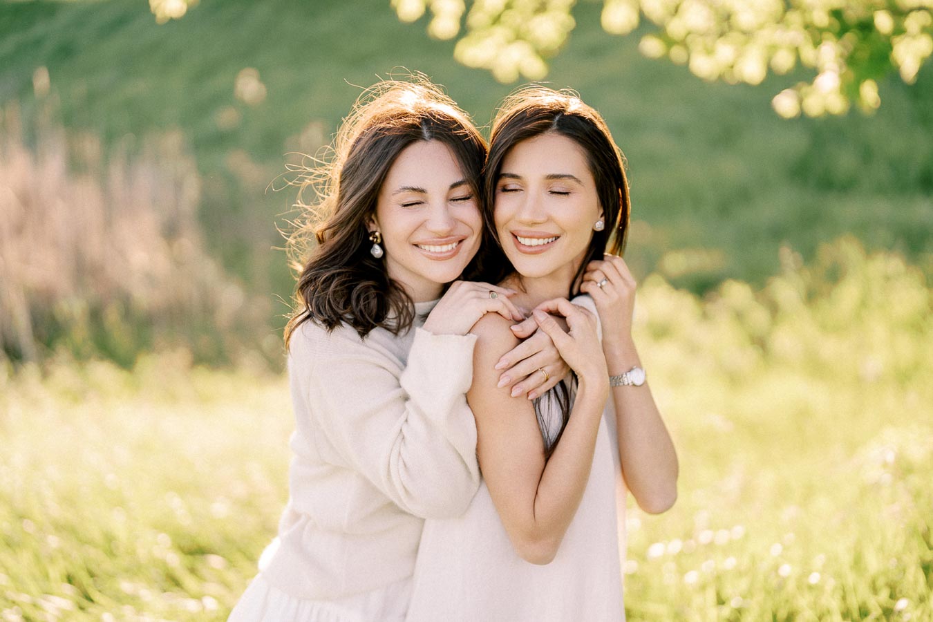 Two women smiling and embracing each other in a sunny, green outdoor setting.