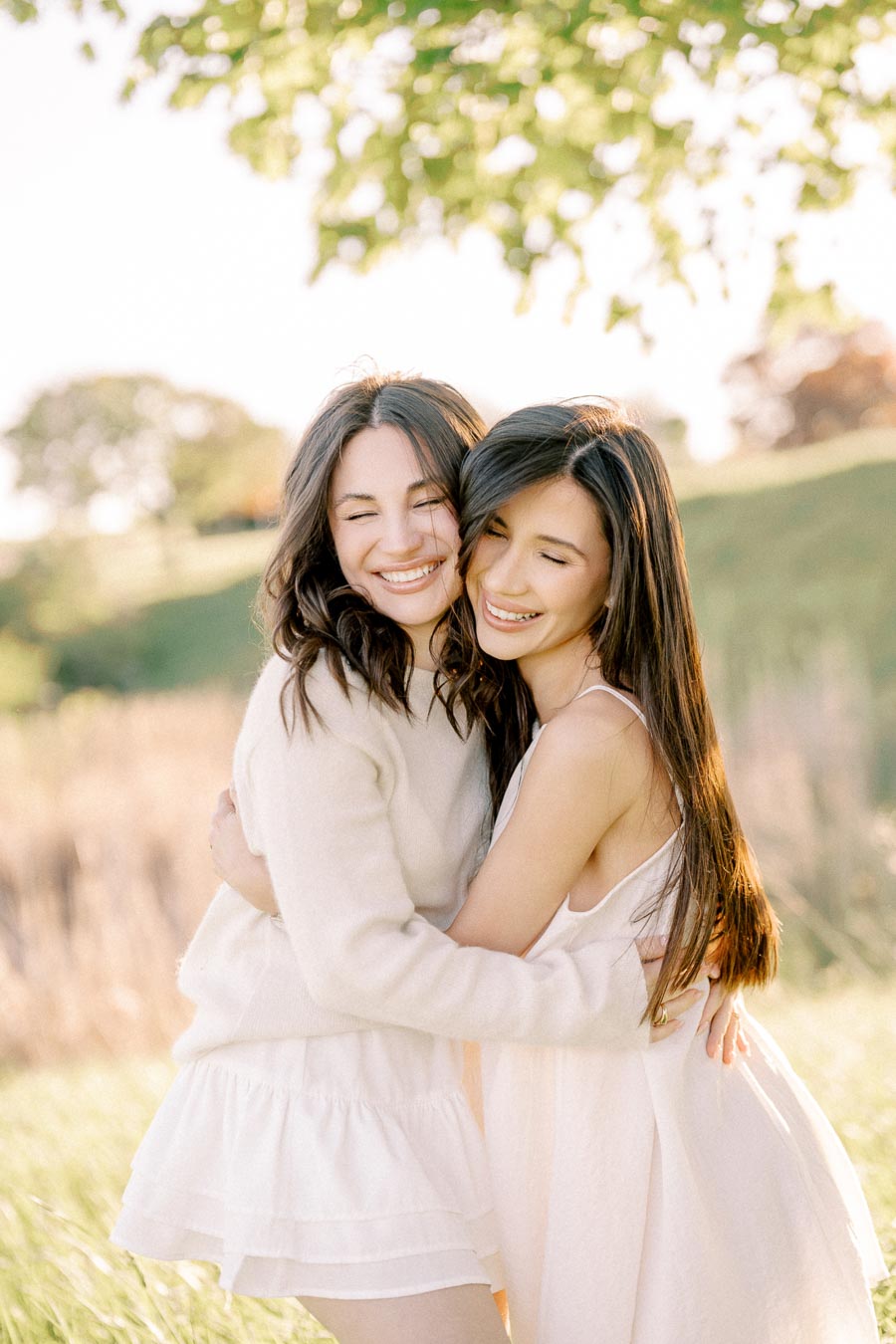 Two women embracing and smiling in a sunny outdoor setting, surrounded by greenery. They are wearing light-colored dresses, enjoying a joyful moment in a park.