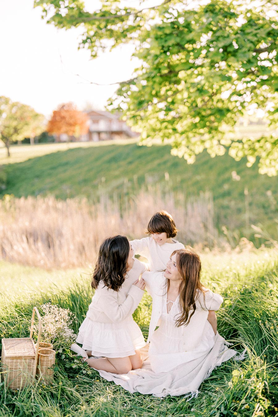 A serene family picnic scene with three people sitting on a lush green lawn under a tree. They are enjoying a sunny day, surrounded by nature, with a wicker basket and flowers nearby.