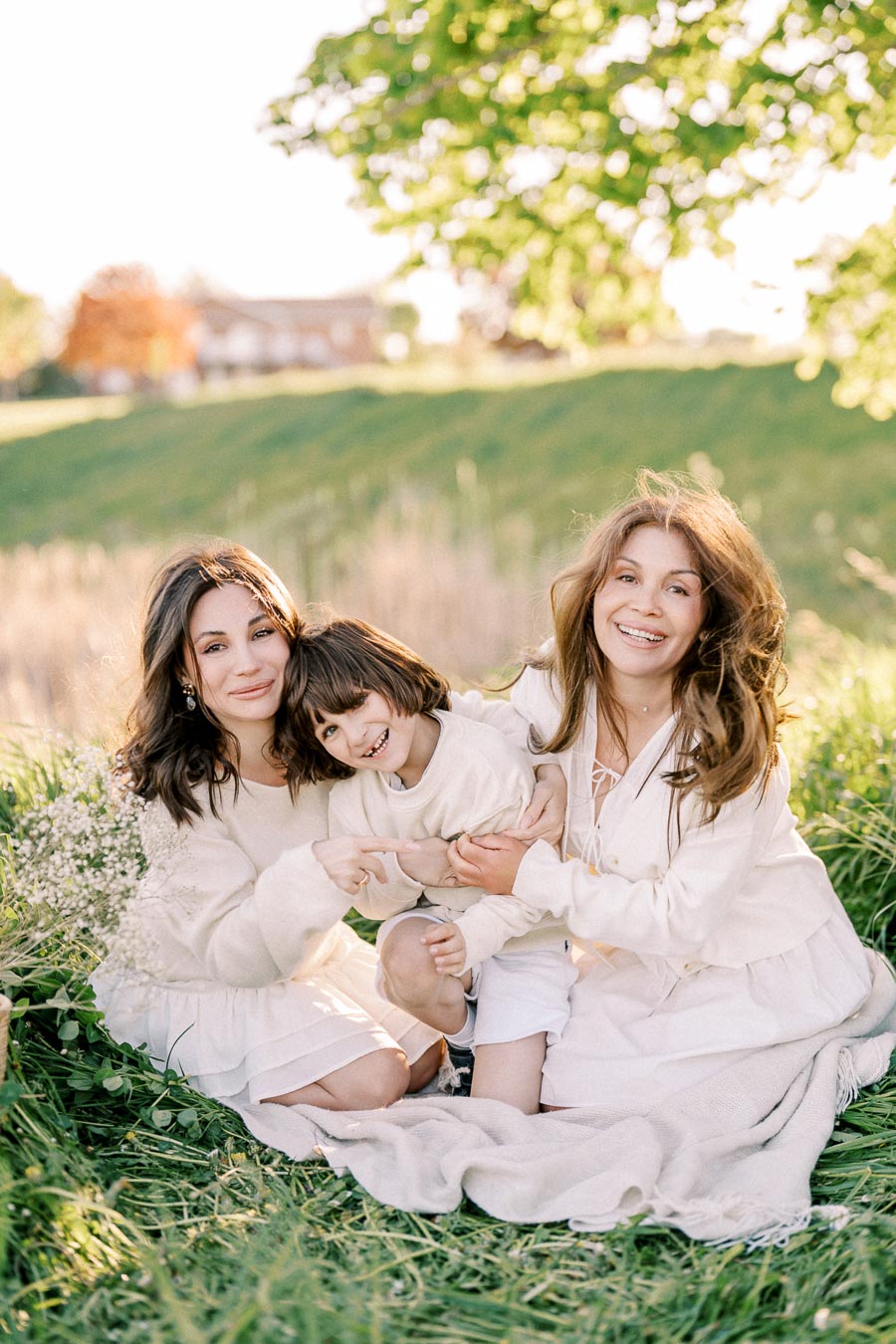 A joyful family sitting on a grassy field during a sunny day, with two women and a child dressed in white, smiling and enjoying the outdoors amidst lush greenery.