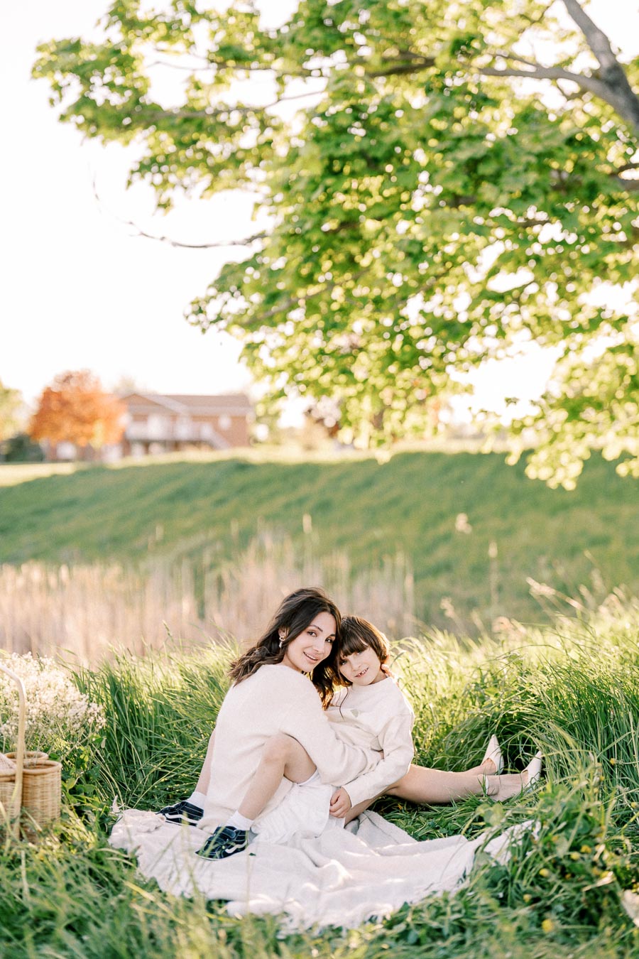 Mother and child enjoying a sunny day outdoors on a picnic blanket, surrounded by lush green grass and trees, with a cozy basket and warm, natural light.