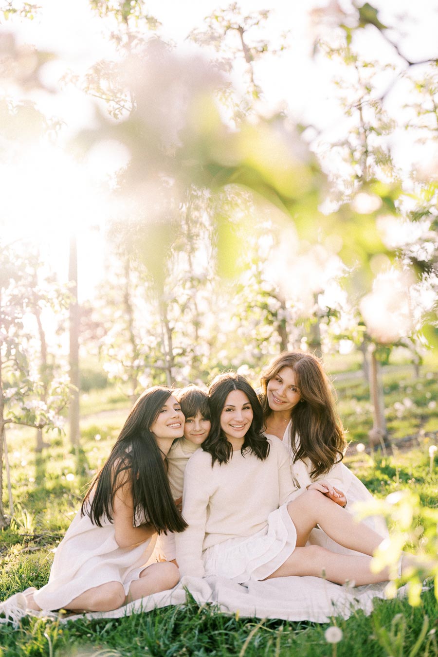 A group of four women sitting on lush green grass in a sunny orchard, wearing light-colored outfits, surrounded by blooming trees and soft natural light.