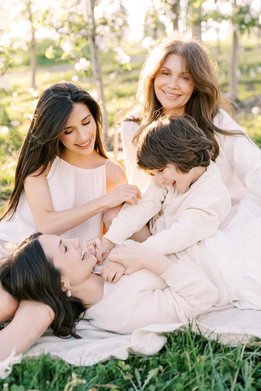 A happy family enjoying a sunny day outdoors, with three women and a child sitting on grass surrounded by blossoming trees, all wearing light-colored clothing and smiling together.