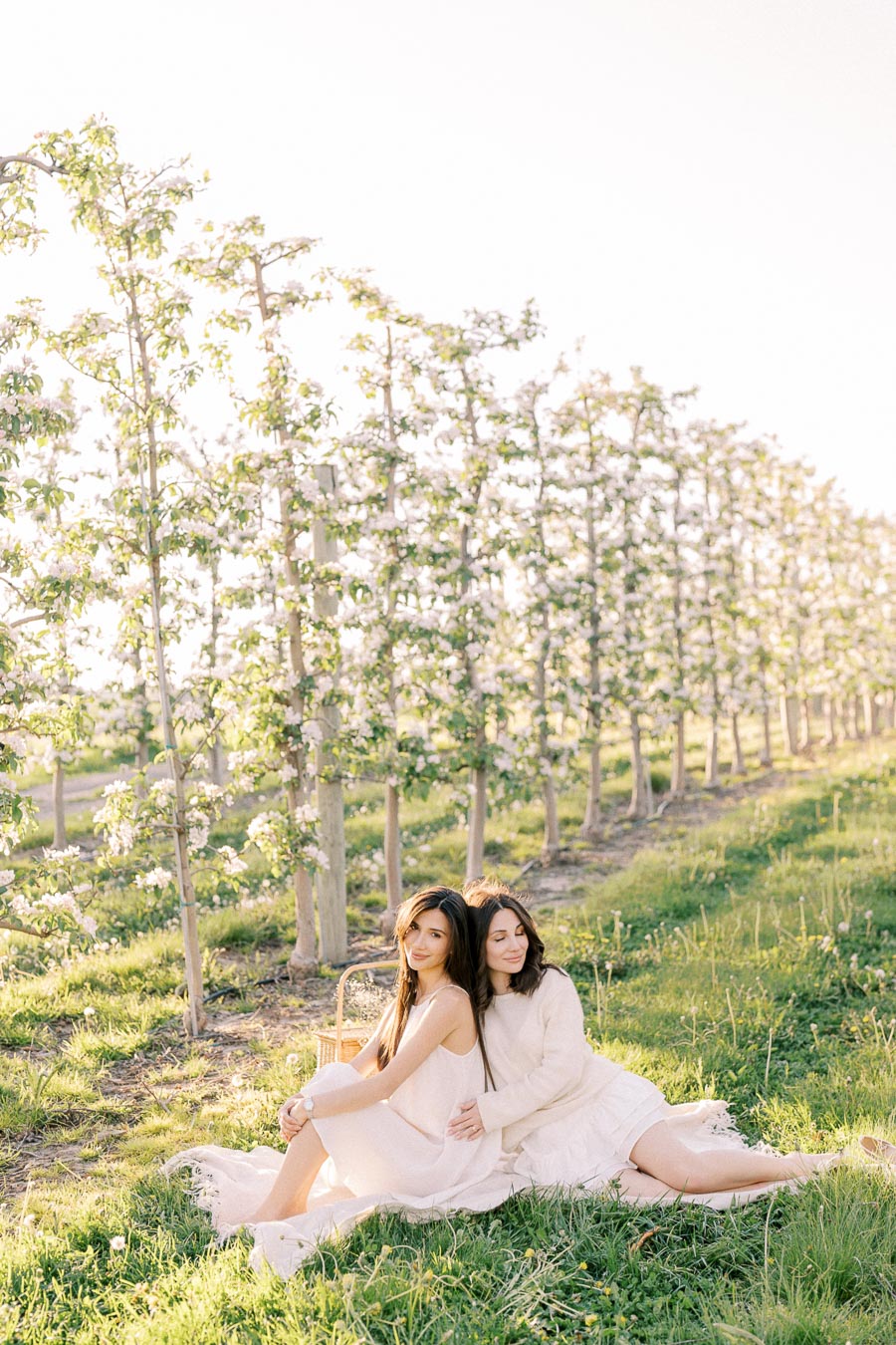 Two women in white dresses sitting on grass in a sunlit orchard, surrounded by blooming apple trees.
