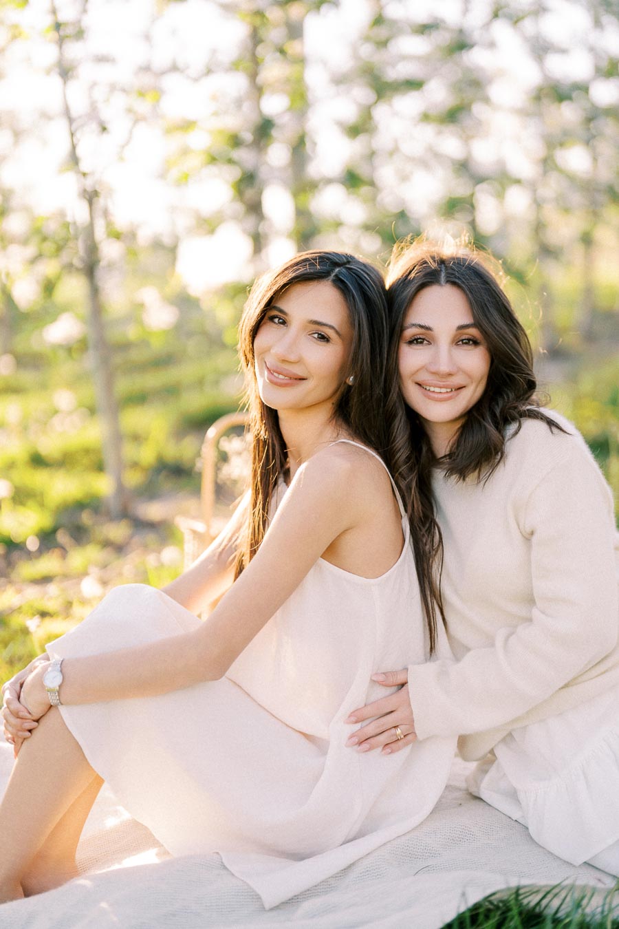 Two women sitting outdoors, smiling and posing in a sunlit garden setting. The woman on the left is wearing a sleeveless dress, while the woman on the right wears a long-sleeved top. The scene is warm and tranquil, with soft natural light and blurred greenery in the background.