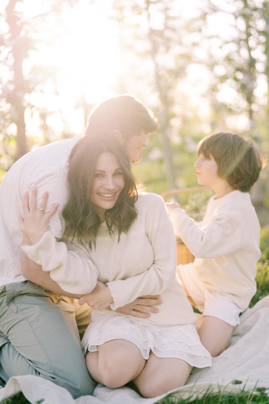Happy family enjoying a sunny day outdoors, with a smiling woman embracing a man, and a child with a basket in the background, surrounded by lush greenery and sunlight.