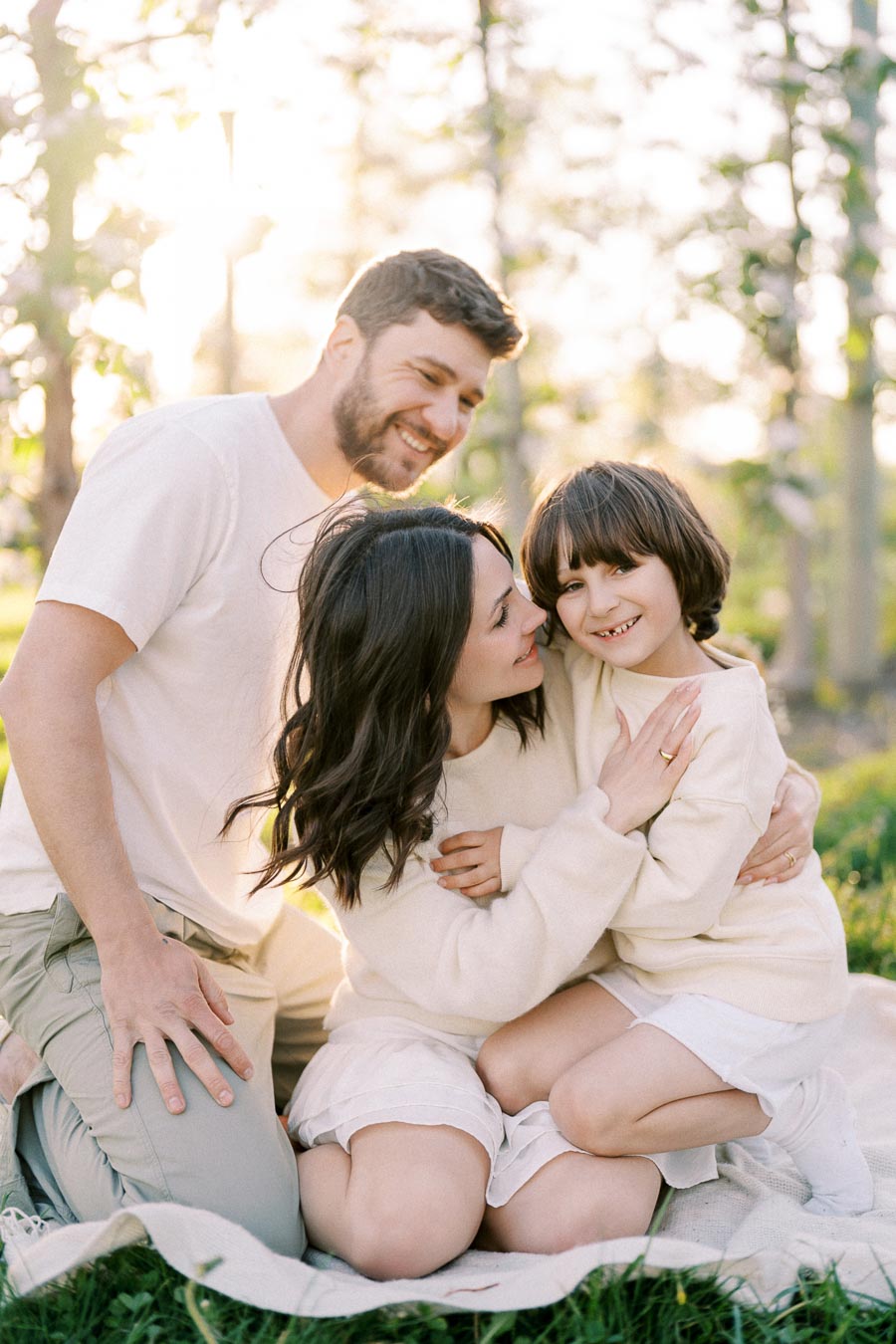 A happy family enjoying a sunny day outdoors, sitting on a blanket in a park, with trees and sunlight in the background. The parents and child are smiling and embracing, dressed in casual light-colored clothing.