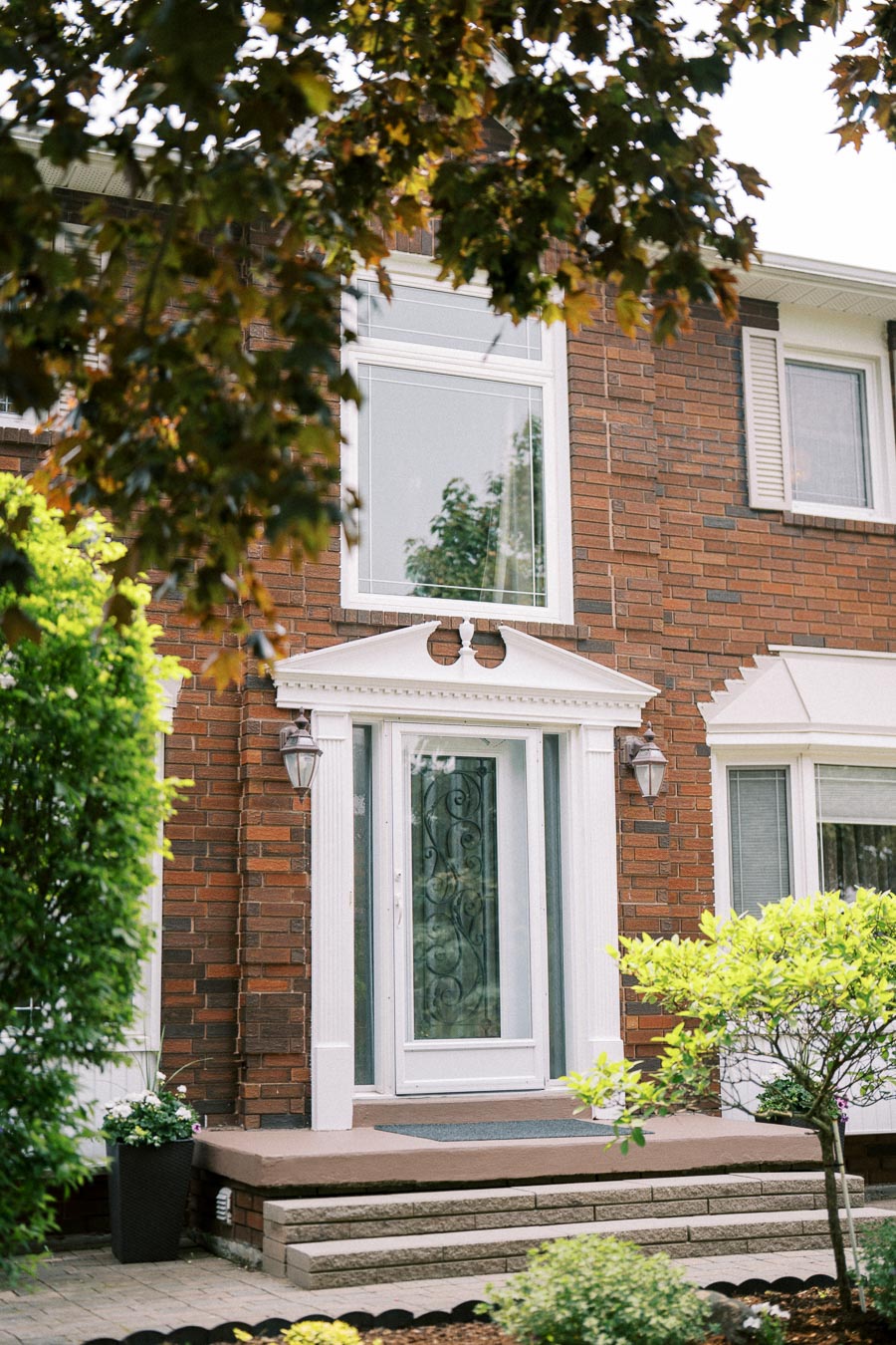 Charming brick house entrance with white door, ornate glass design, and greenery, featuring lush foliage and a potted plant, creating a welcoming atmosphere.